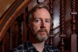 A man with a trim beard, in a dark blue plaid shirt, stands with arms crossed in front of a carved wooden bannister.
