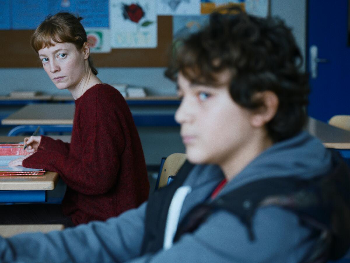 A teacher in a dark red sweater looks quizzically at a young teen student sitting near her.