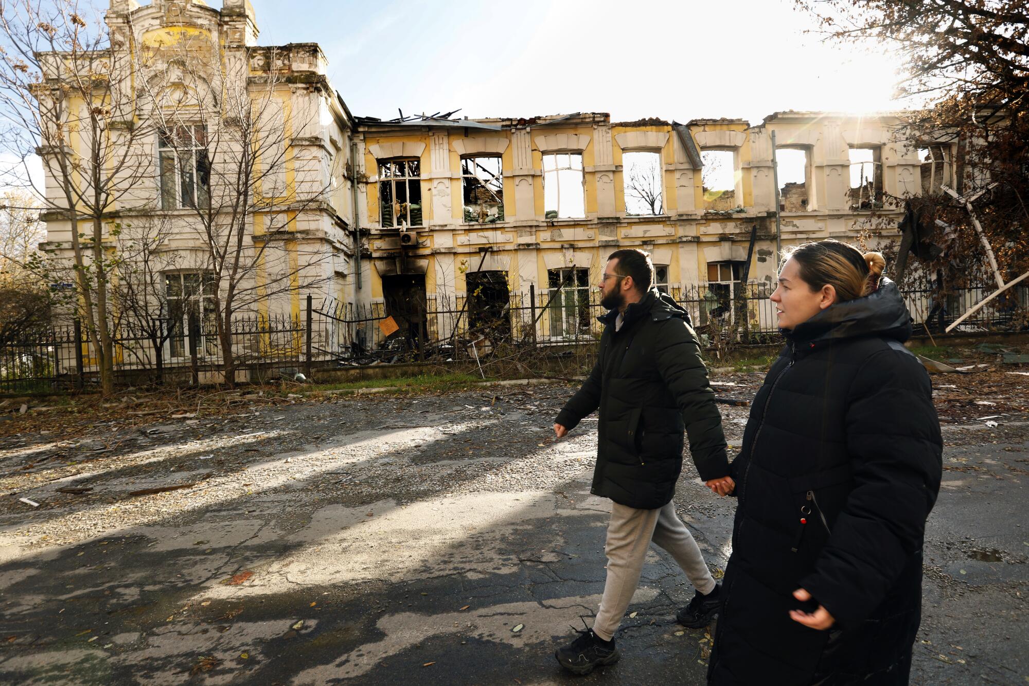 A bearded man, left, and a woman, both in dark winter jackets, hold hands as they walk past a destroyed building 