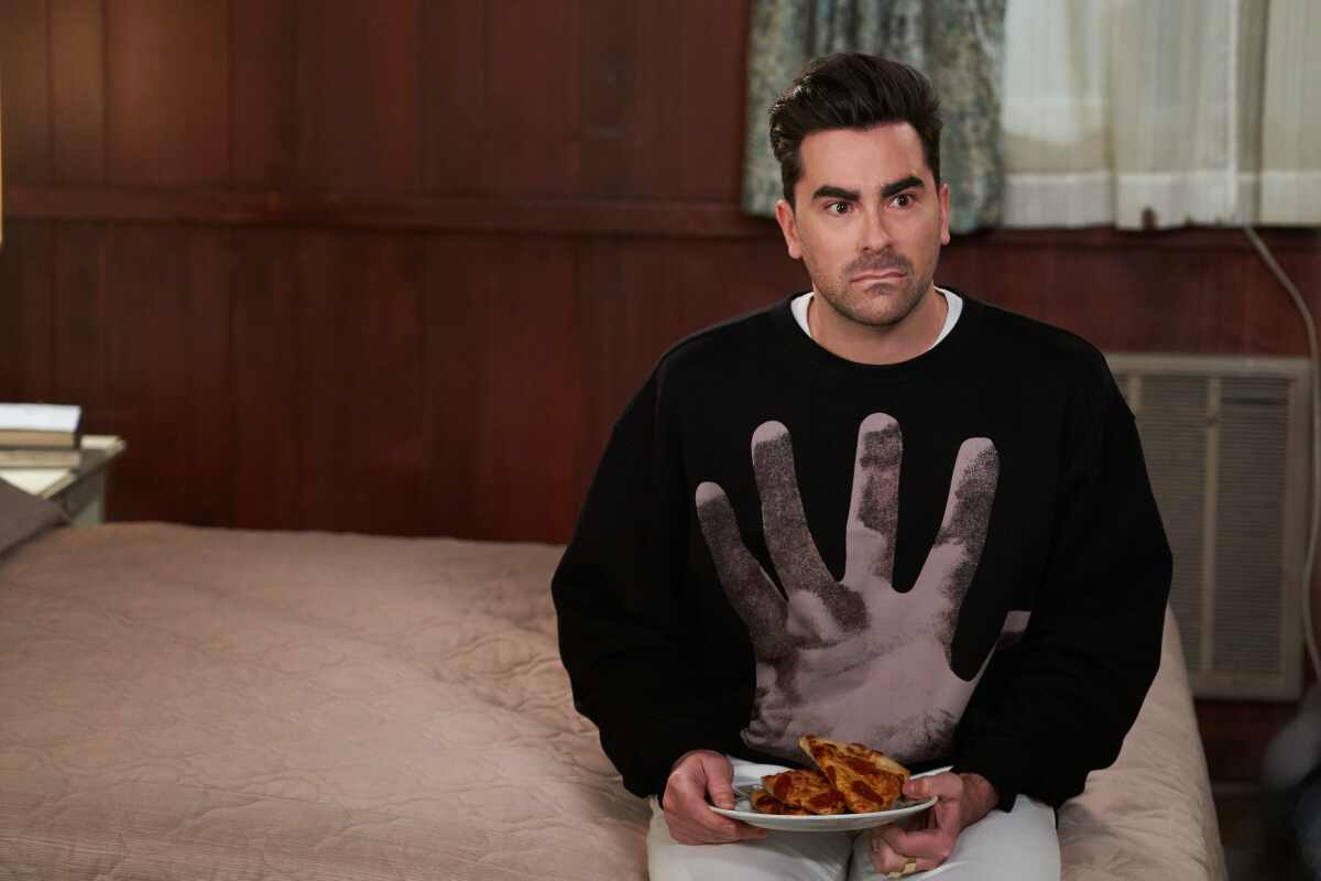 Dan Levy sits on a hotel bed with a plate of food in his role as David Rose on "Schitt's Creek."