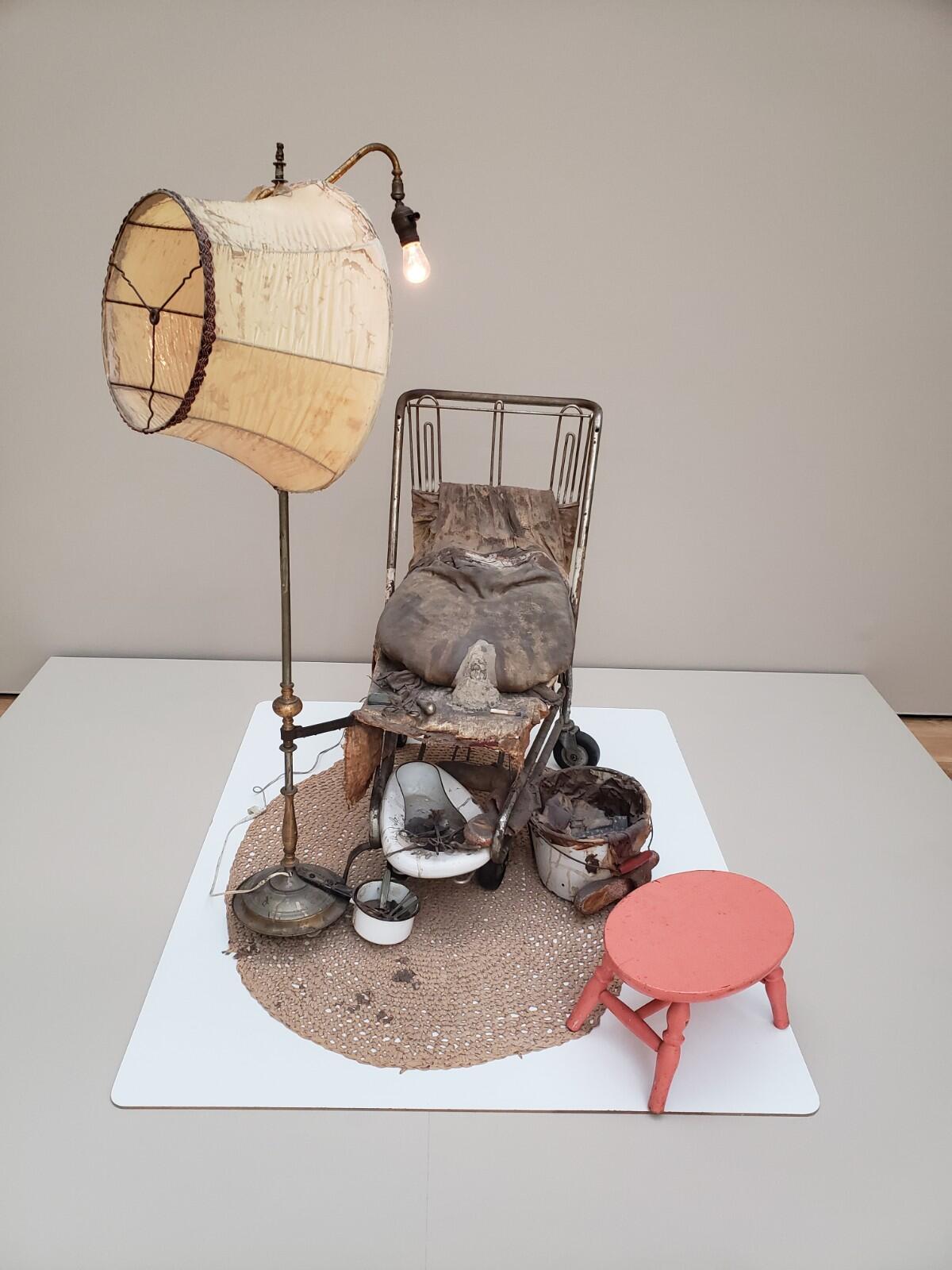 A lamp with its shade pushed off stands beside a chair with a pack of cement. Beneath is a pile of medical implements.