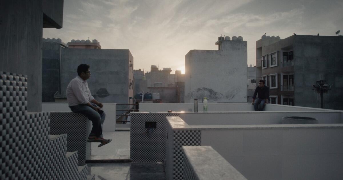 Two men sit across an urban rooftop from each other.
