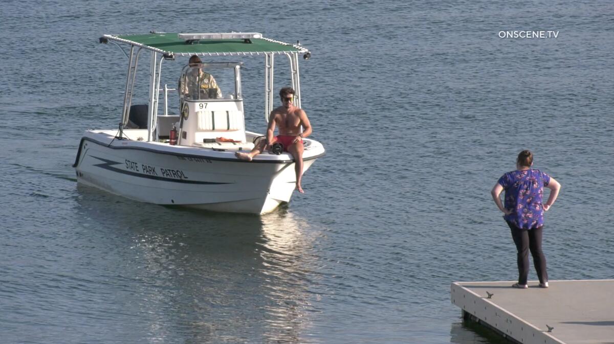A man in swim trunks sits on the front of small boat as a woman watches from a dock. 