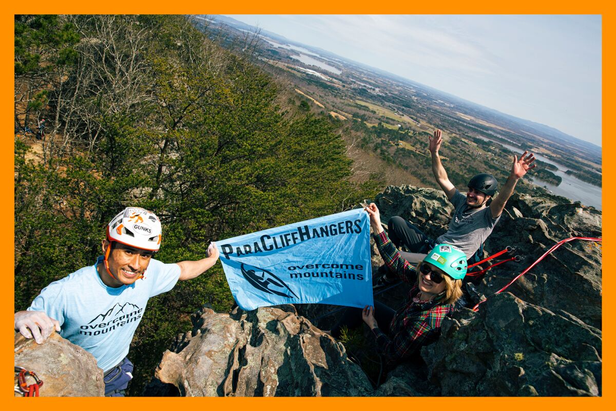 A group of climbers hold a ParaCliffHangers flag at the top of a mountain.