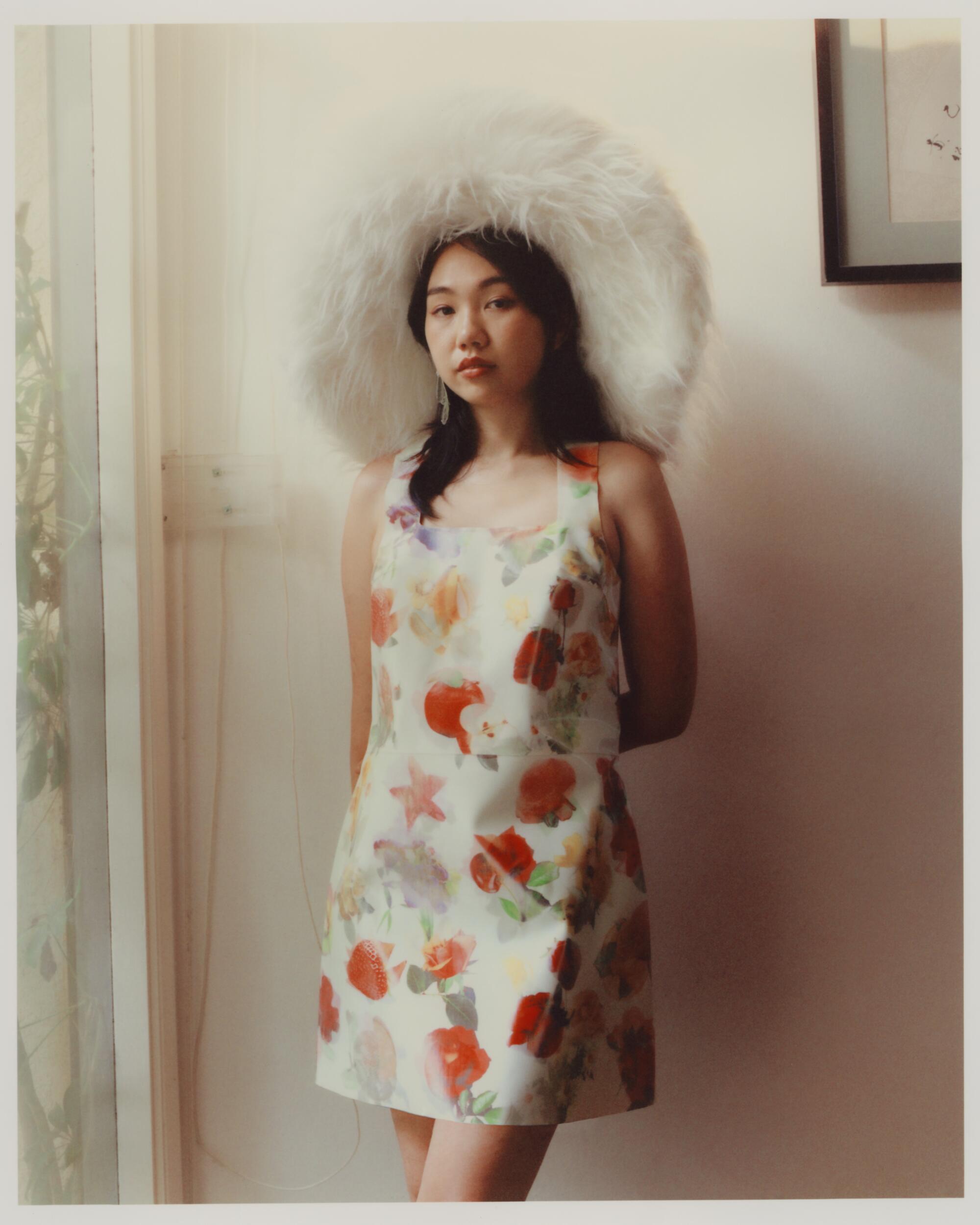 A woman stands against a light-colored wall wearing a big fluffy hat and a shift dress printed with fruits.