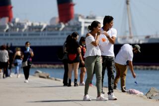 LONG BEACH, CA - JULY 18: People walk along the board walk with the Queen Mary in the background near Shoreline Village on Sunday, July 18, 2021 in Long Beach, CA. Los Angeles County on Sunday began requiring people to wear masks in indoor public places. (Gary Coronado / Los Angeles Times)