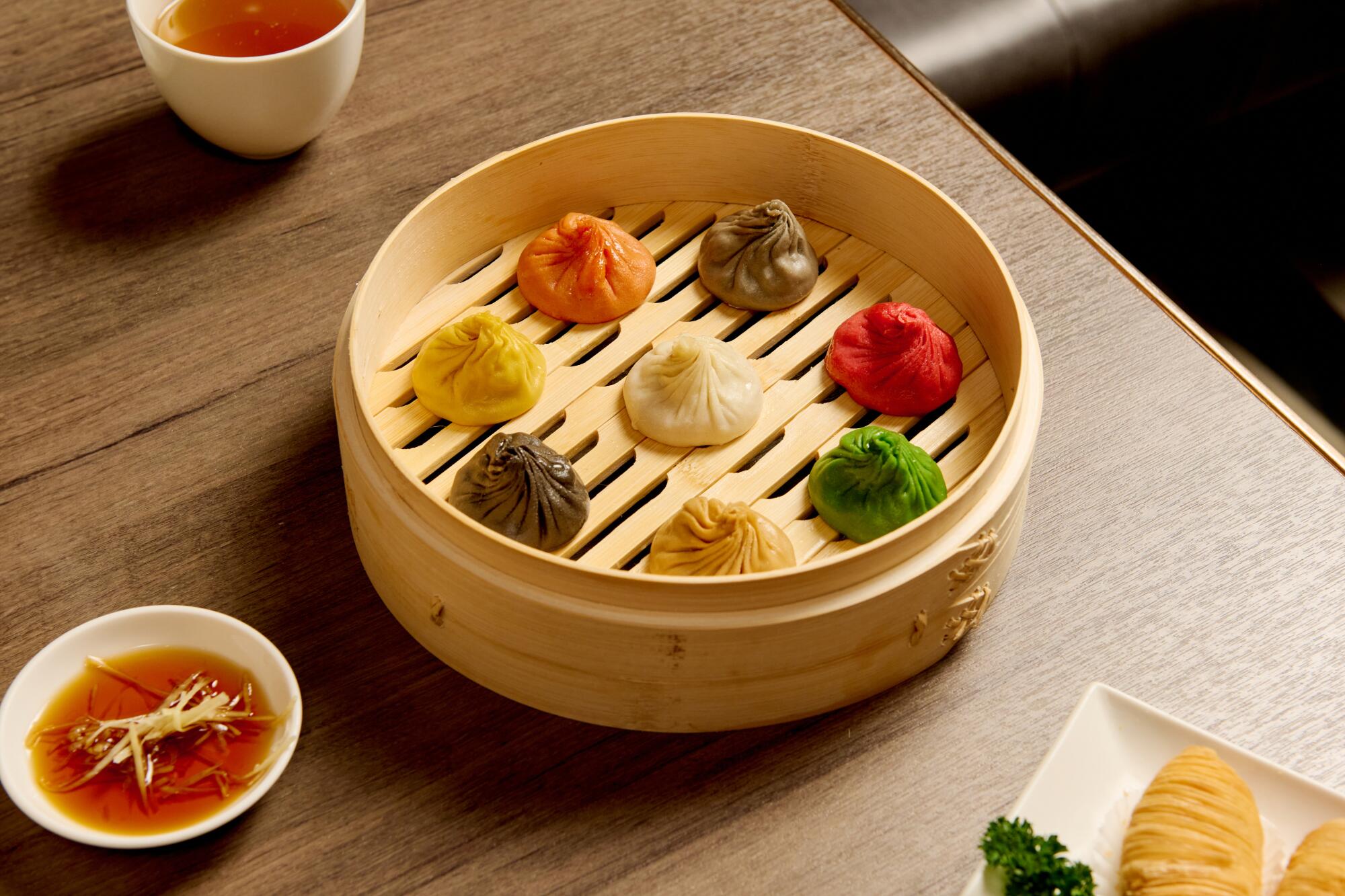 A rainbow of soup dumplings in a steamer basket with small dishes of sauces