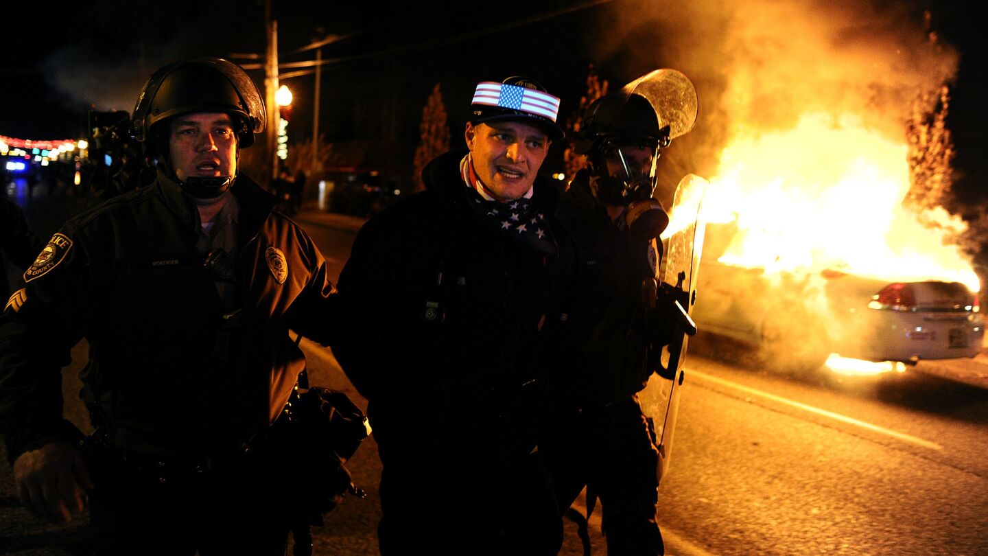 A protestor is arrested as police escort him by a burning police car .
