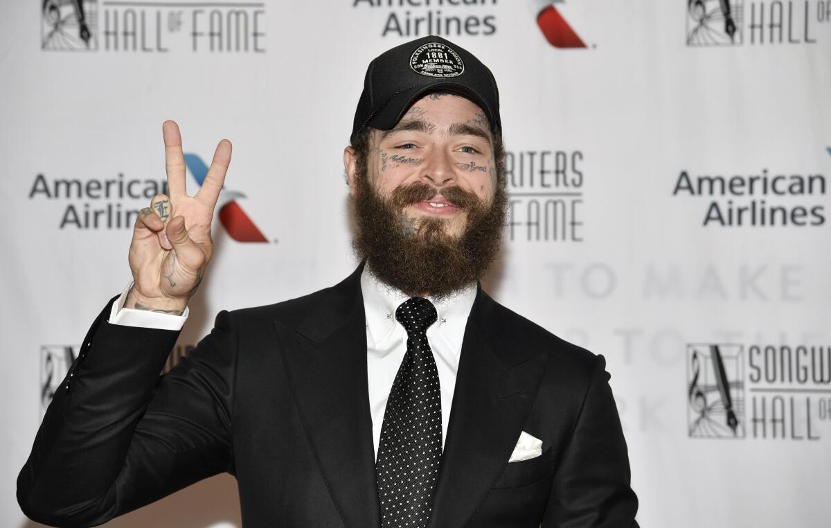 Post Malone flashes a peace sign as he poses in a black suit, white shirt, tie and a black ballcap
