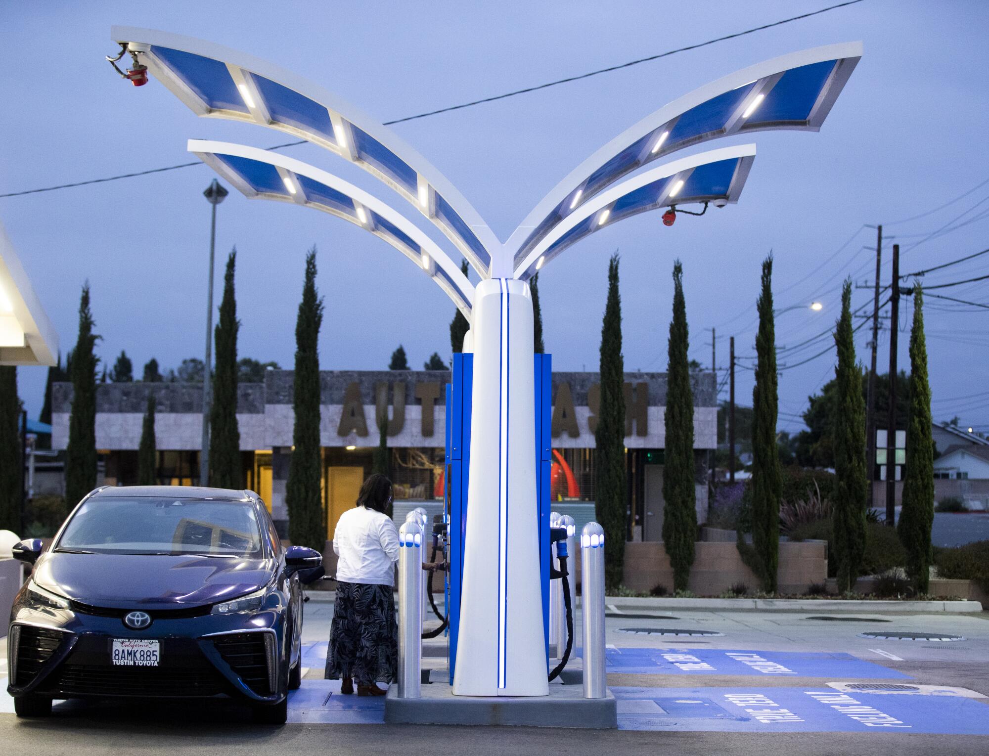 A person stands next to his vehicle under a dark gray sky.  They are in a Y-shaped hydrogen fuel pump.