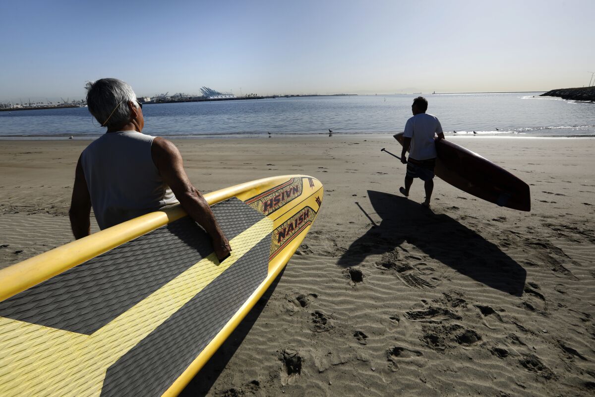 Two men carry paddle boards on a beach toward the ocean.