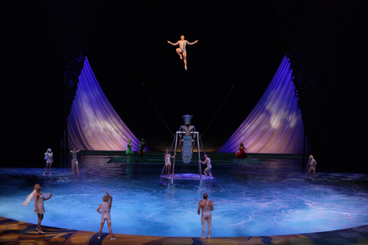 An acrobat is suspended above a pool. Other performers are alongside it and on platforms in the water.