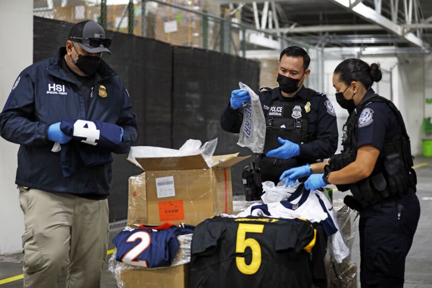 Los Angeles, California-Feb. 4, 2022-U.S. Customs and Border Protection officers demonstrate the opening of boxes of counterfeit goods the agency has recovered during a press conference on Feb. 4, 2022. Ahead of the Super Bowl, U.S. Customs and Border Protection (CBP) and U.S. Homeland Security Investigations(HSI) in partnership with other federal, state and local partners form a united front and are conducting enforcement operations to combat the trafficking of NFL counterfeits. They display some of the counterfeit NFL jerseys, caps, championship rings and a fake Lombardy Trophy have recovered recently. (Carolyn Cole / Los Angeles Times)
