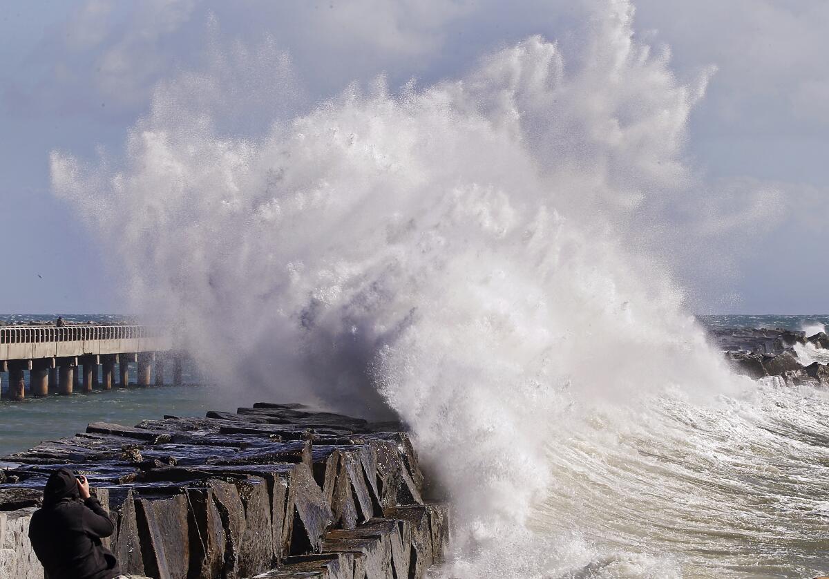 A large wave captures into a ocean breakwater. In the background is a pier and blue sky.