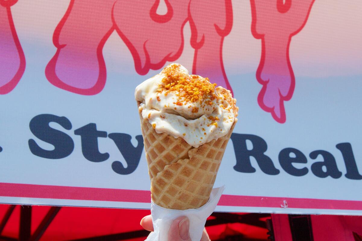 A hand holds a waffle cone of honeycomb-topped Creamy Boys New Zealand-style whipped soft serve in front of the stand's sign.