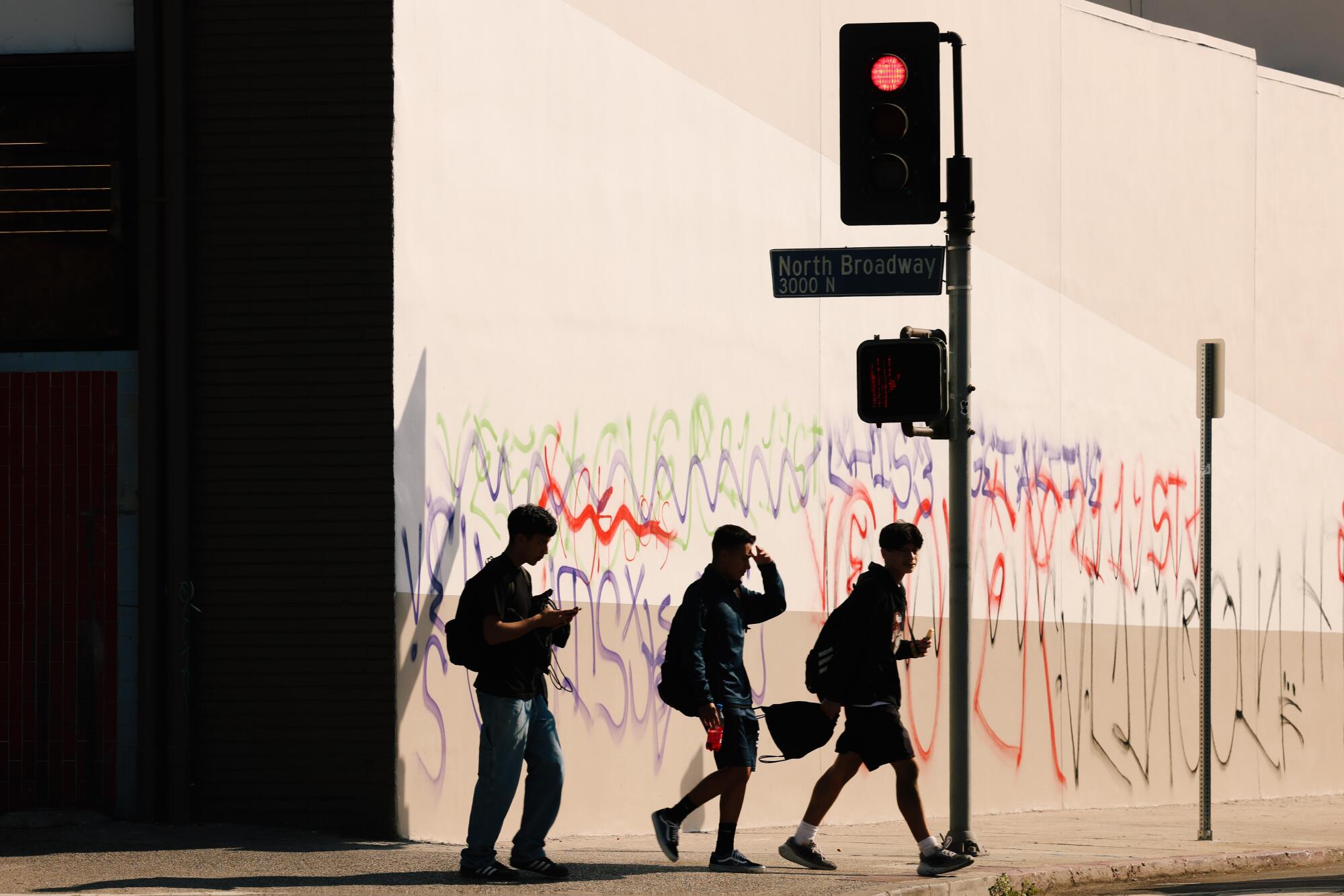 Three young men walk toward a crosswalk, a wall full of colorful graffitti stands behind them. 