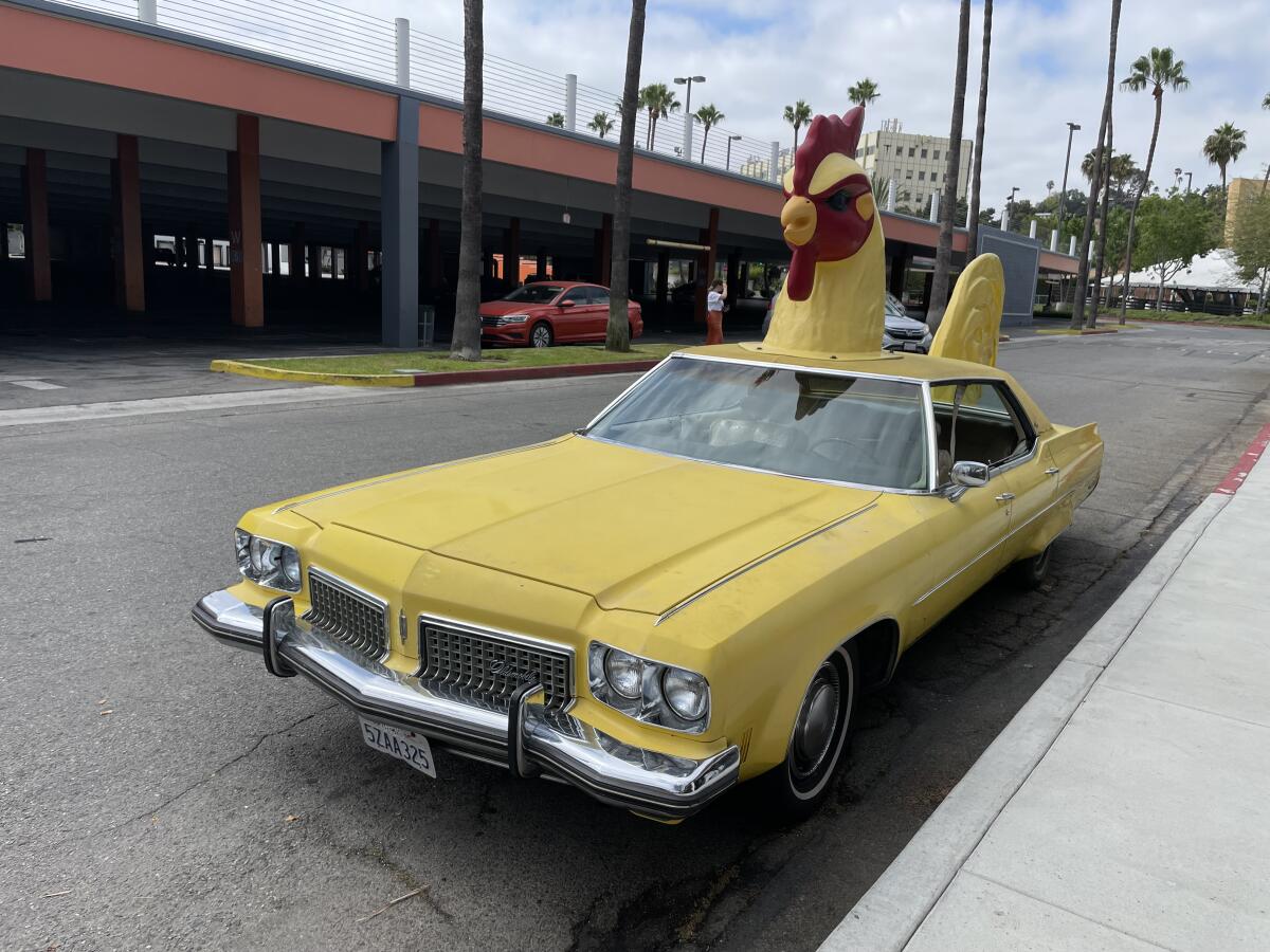 A yellow car with a chicken head on top and tail on the back.