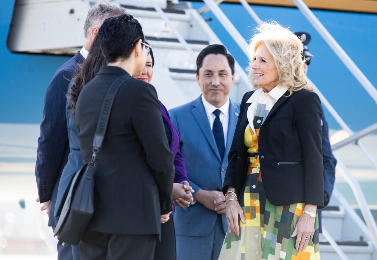 A smiling blond woman stands on a tarmac at the foot of a set of airplane stairs surrounded by people in suits