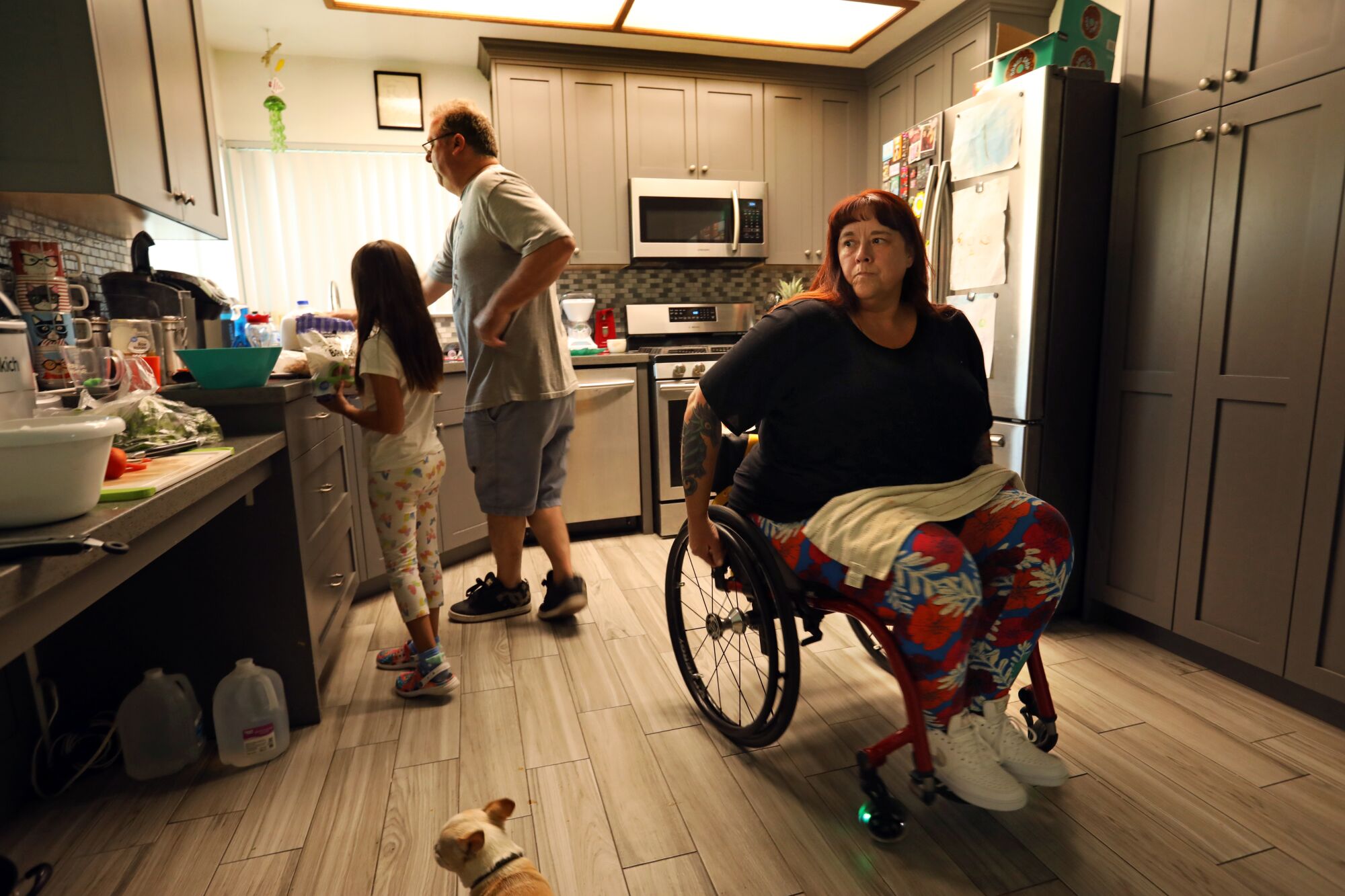 A woman sits in a wheelchair in her kitchen as her husband and granddaughter prepare food at the counter