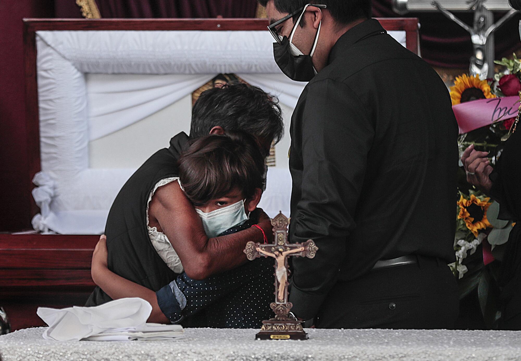 Two people in masks embrace in front of an open casket behind a crucifix as another person stands nearby