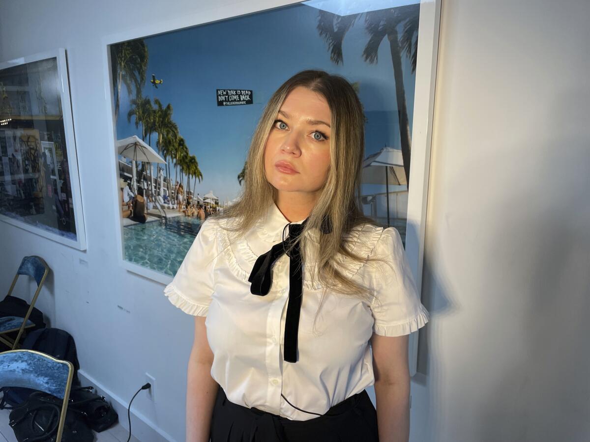 Anna Delvey in a white button down shirt, a black tie and black pants posing in front of a poster with a tropical setting