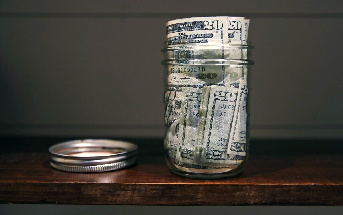 A jar filled with $20 bills sits on a shelf. Its lid is to the left.