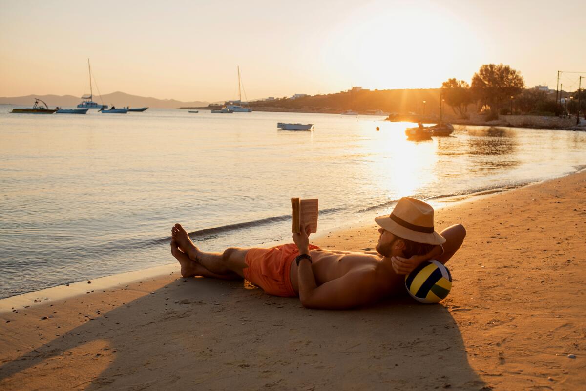 A man lies on the sand at the beach, reading a book.