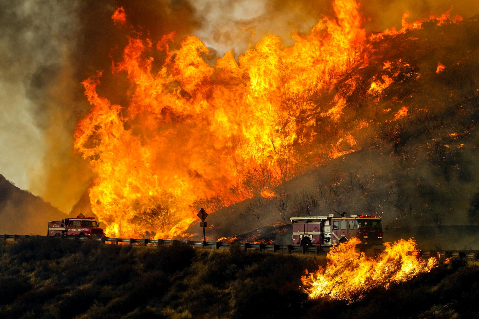 Fire trucks drive past a wall of flame