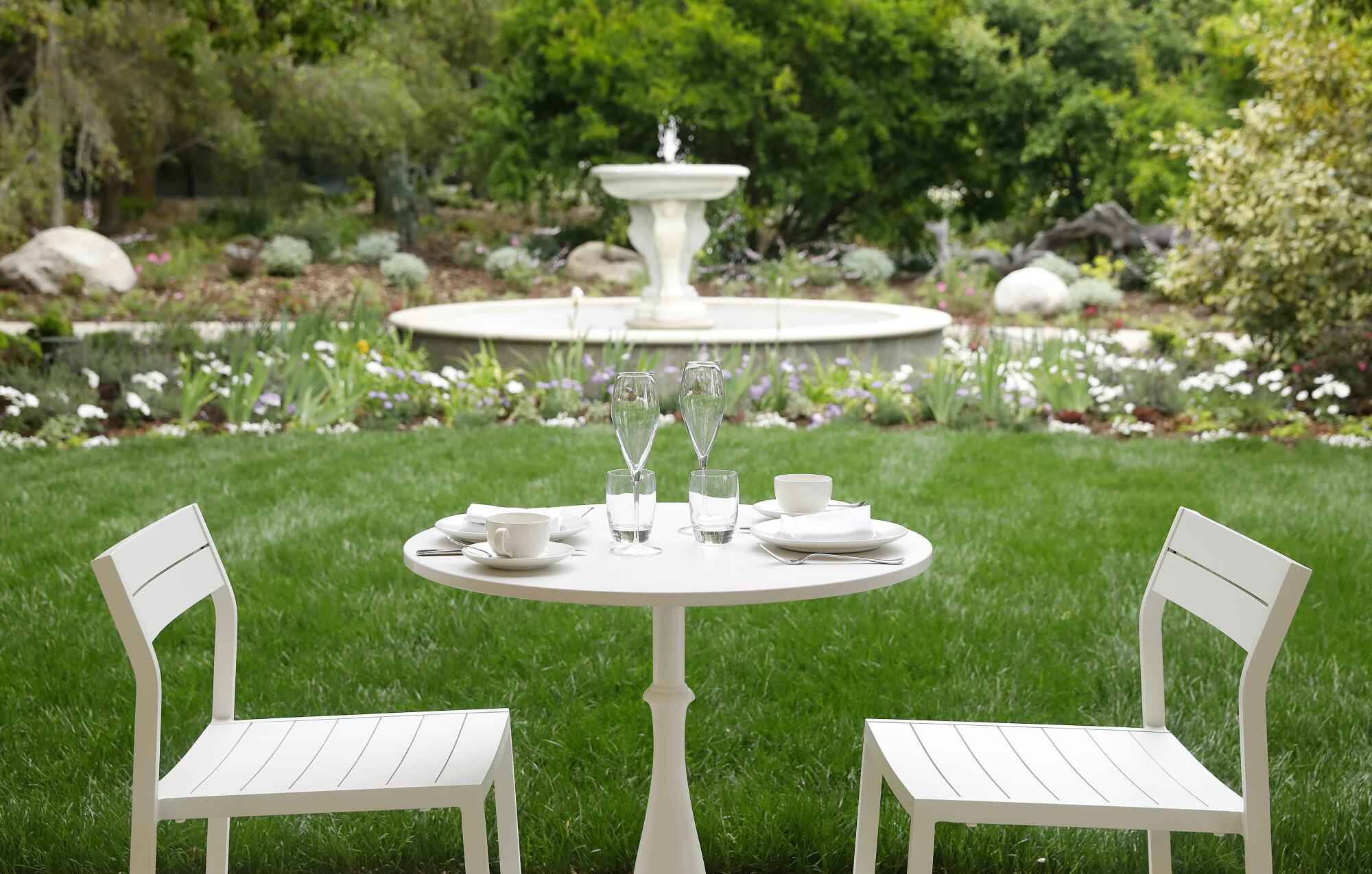 A white table with two chairs, with a garden fountain beyond.