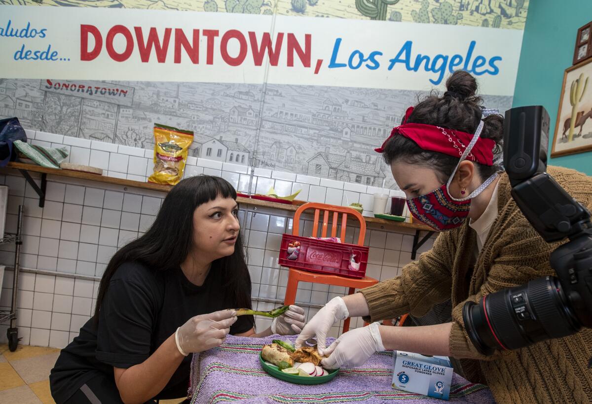 A seated woman, left, looks at a woman in a mask and gloves adjusting a plate of food on a table
