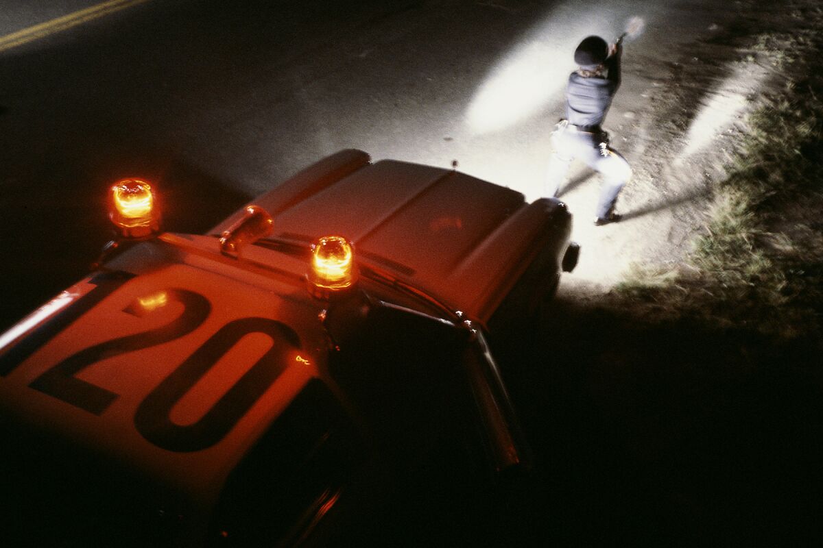 A police officer stands and points a gun in front of a police car's headlights.
