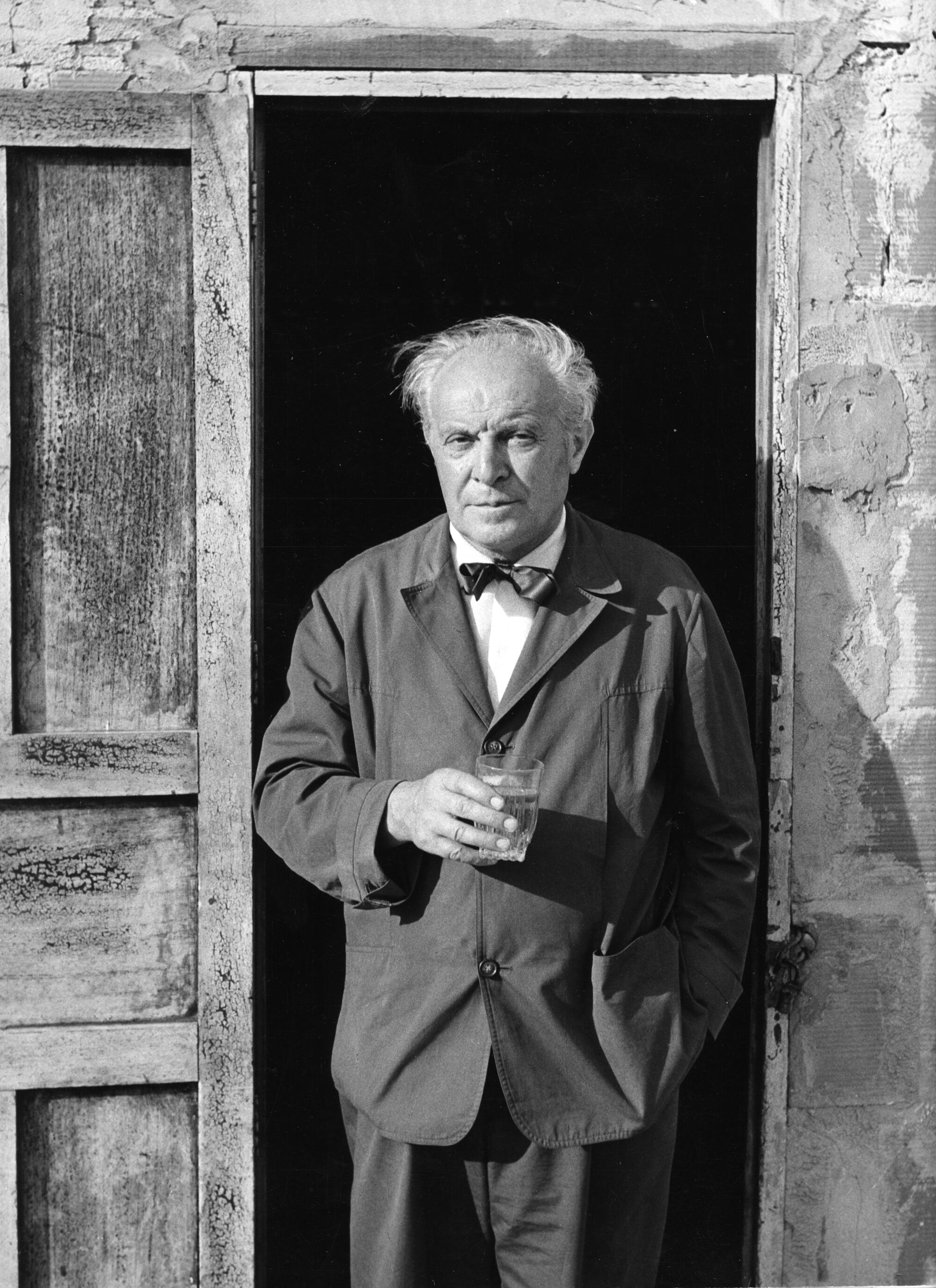 Gio Ponti, in a workman's jacket and bow tie, stands in the doorway of a rough-hewn building
