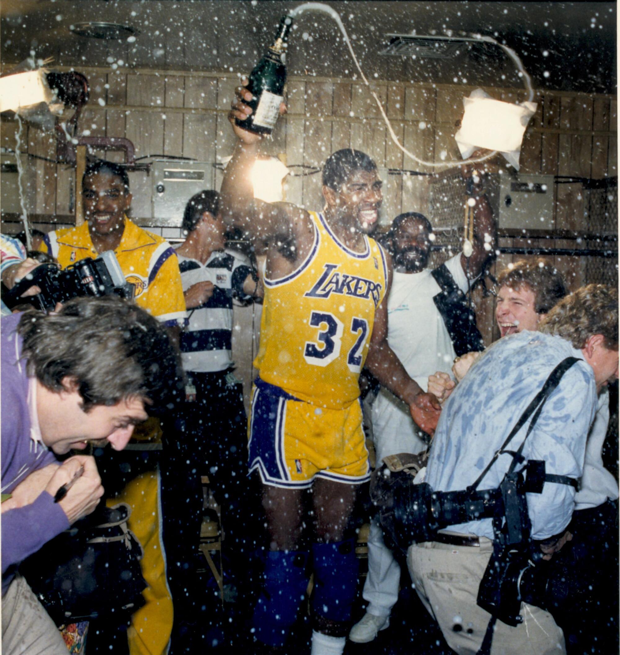 Magic Johnson in a gold Lakers jersey splashes champagne from a bottle around a locker room with players and photographers.