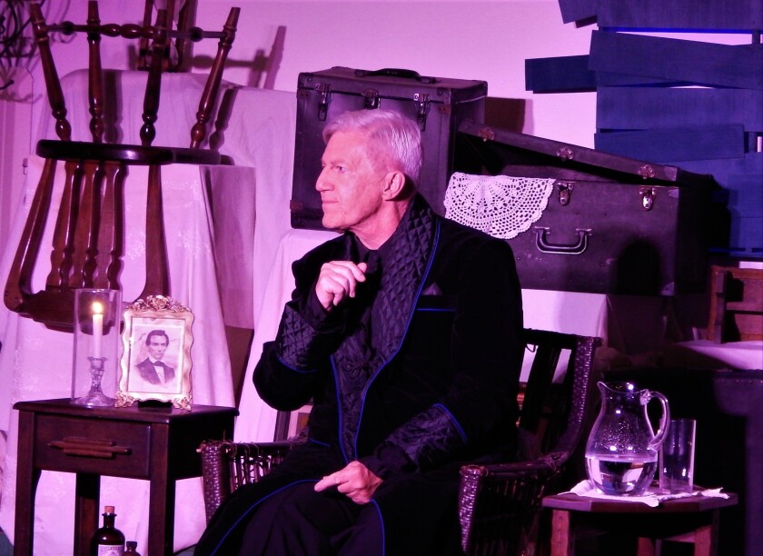 Actor Tom Dugan sits among antiques, wearing a black dressing robe.