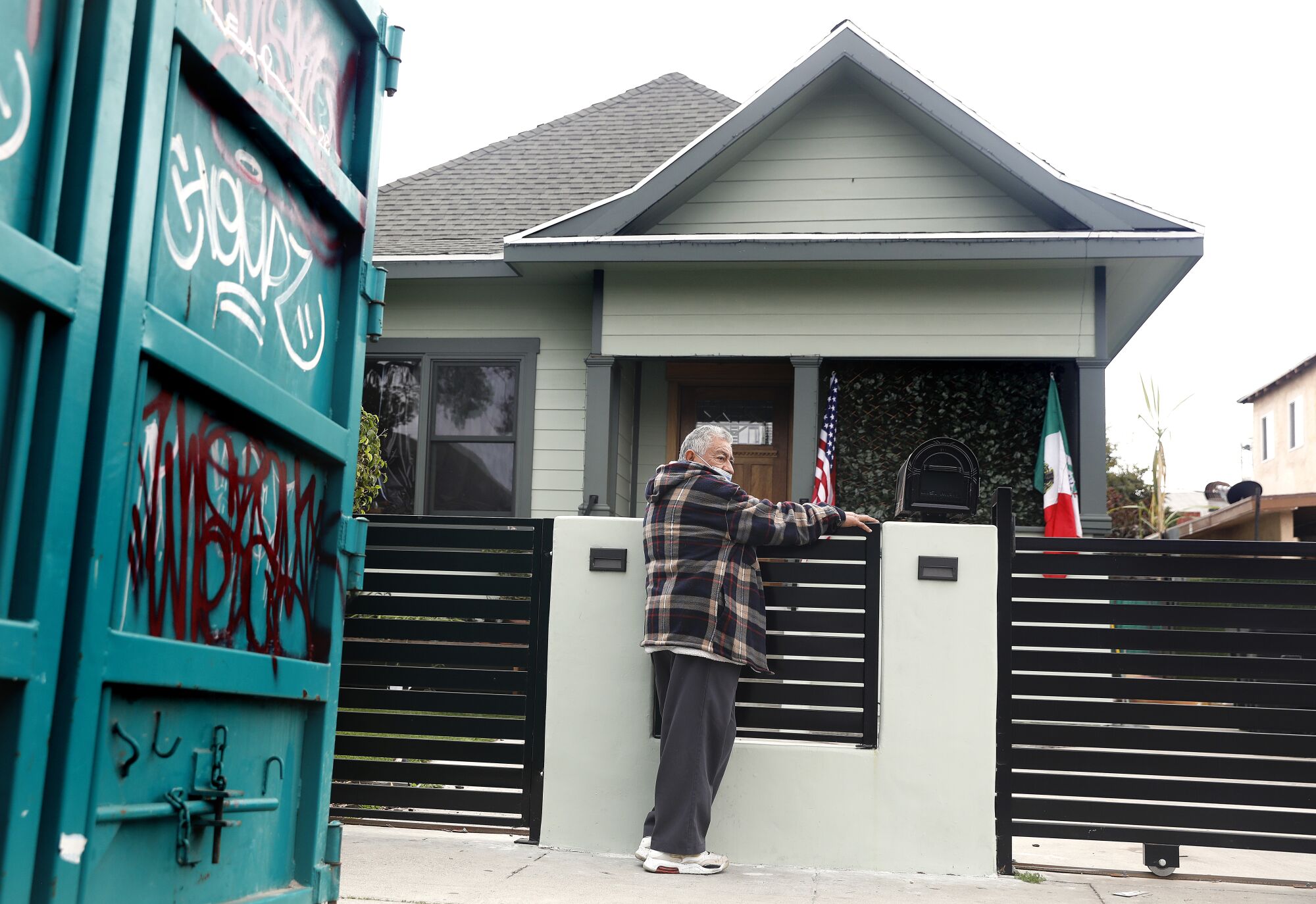 A man stands outside a house displaying U.S. and Mexican flags.