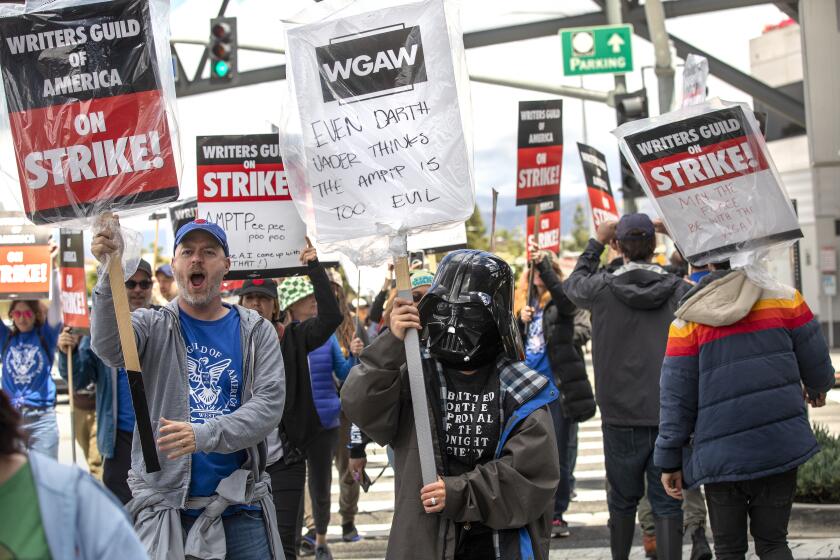 UNIVERSAL CITY, CA-MAY 4, 2023: Joe Henderson, left, a writer and show runner, and Montserrat Luna, a writer, wearing a Darth Vader helmet, strike with other members of the Writers Guild of America outside of NBC/Universal Studios in Universal City. (Mel Melcon / Los Angeles Times)