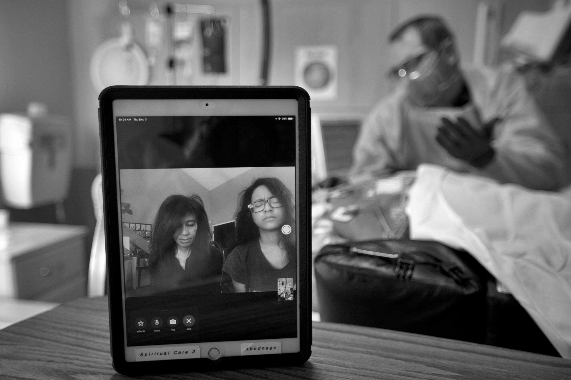 Two people chat by video on an iPad as Chaplain Kevin, at right in background, prays over a patient