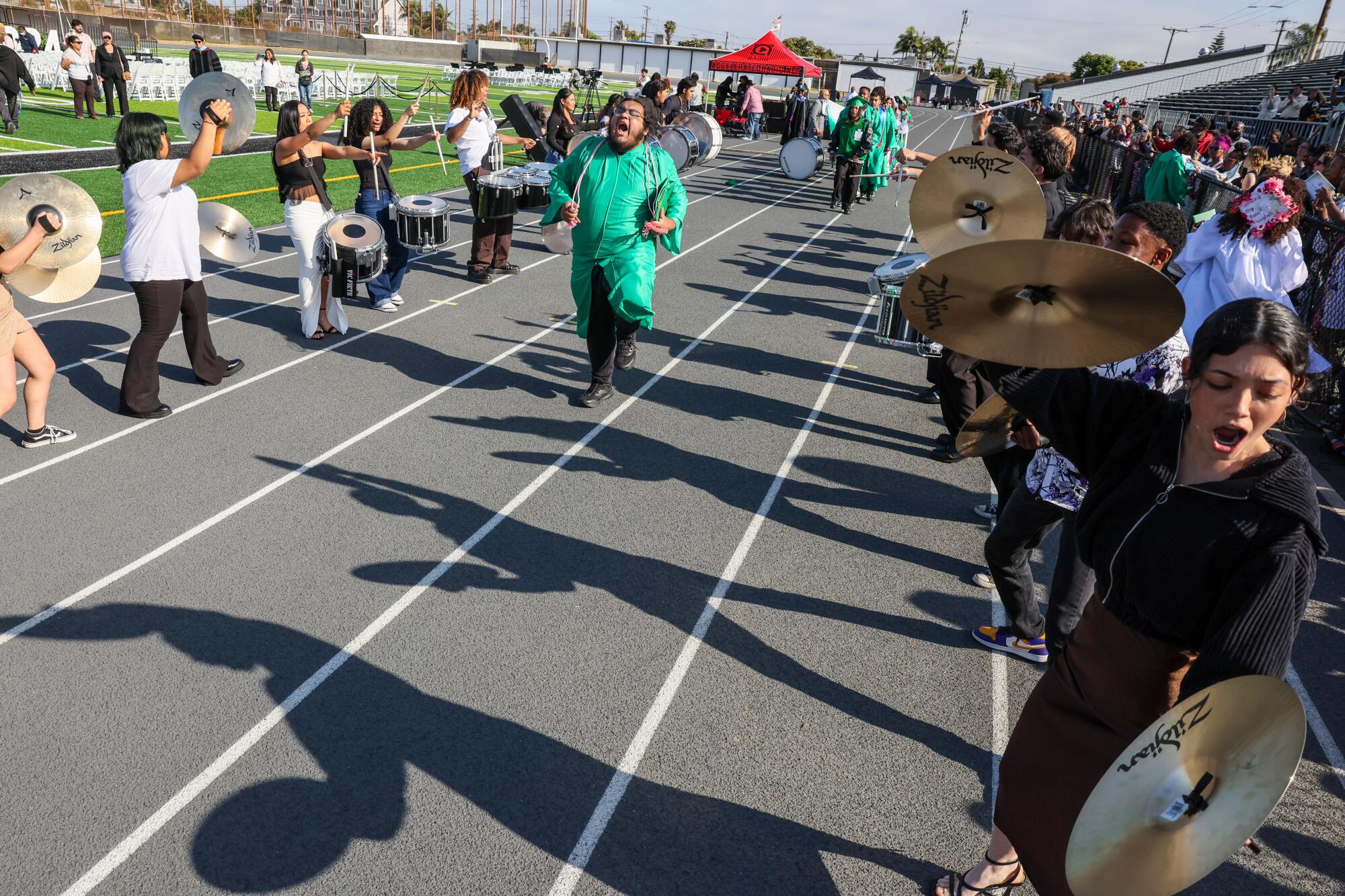 A person in a green robe runs down a track lane while band members play on both sides.