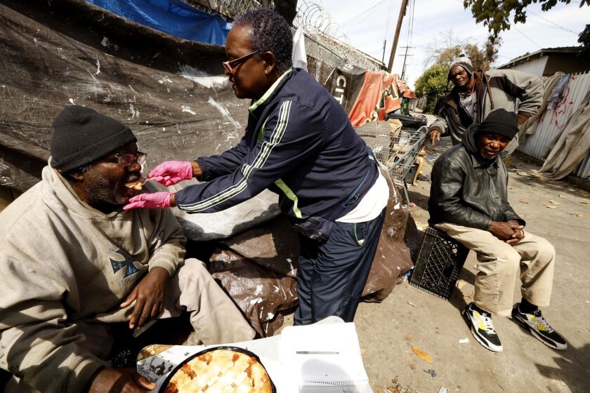LOS ANGELES, CA - MARCH 18, 2018 - Rose Rios, 70, with Cover the Homeless Ministry, center, feeds some pie to Donald Shields, 59, who is legally blind and living in an alley in South Los Angeles on March 18, 2018. He was being taken care of by another pair of homeless men, childhood friends Wayne Robinson, 58, right, and Daniel Murray, 59, background, who were camping next to him. Rose Rios, 70, goes into the worst parts of South Los Angeles to give people money, blankets, clothes and food for her Cover the Homeless Ministry, an organization she started a decade ago. She runs it by herself out of her home, with little to no help from others. (Genaro Molina / Los Angeles Times)