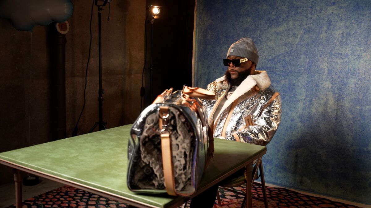 man in shiny silver jacket and sunglasses sits at a table with a designer duffel bag on it