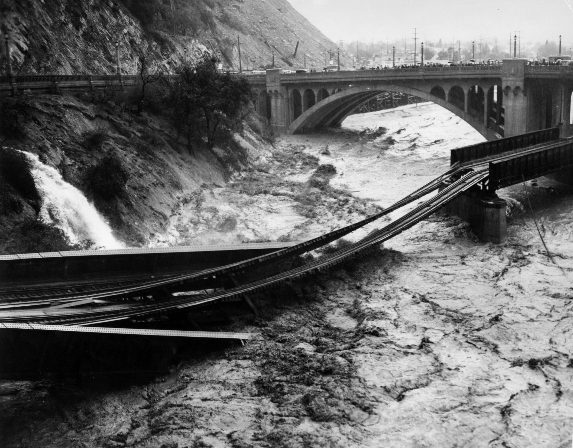 Remnants of a destroyed railroad bridge are suspended over a raging river.