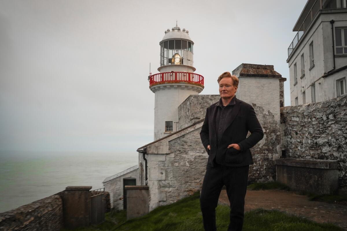 Conan O'Brien, wearing all black, stands in front of a lighthouse.