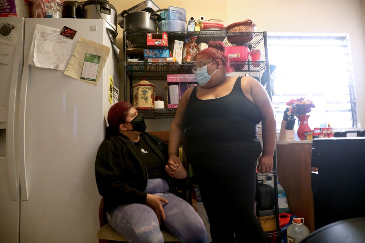 Two women, one seated, wear face masks as they hold hands near a refrigerator in a cramped apartment.