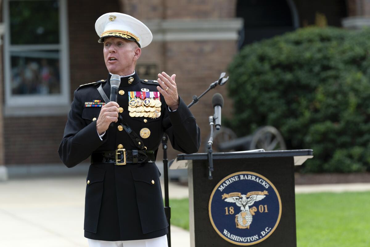 A decorated officer gesturing as he speaks into a microphone outdoors next to a lectern with a U.S. Marine logo.