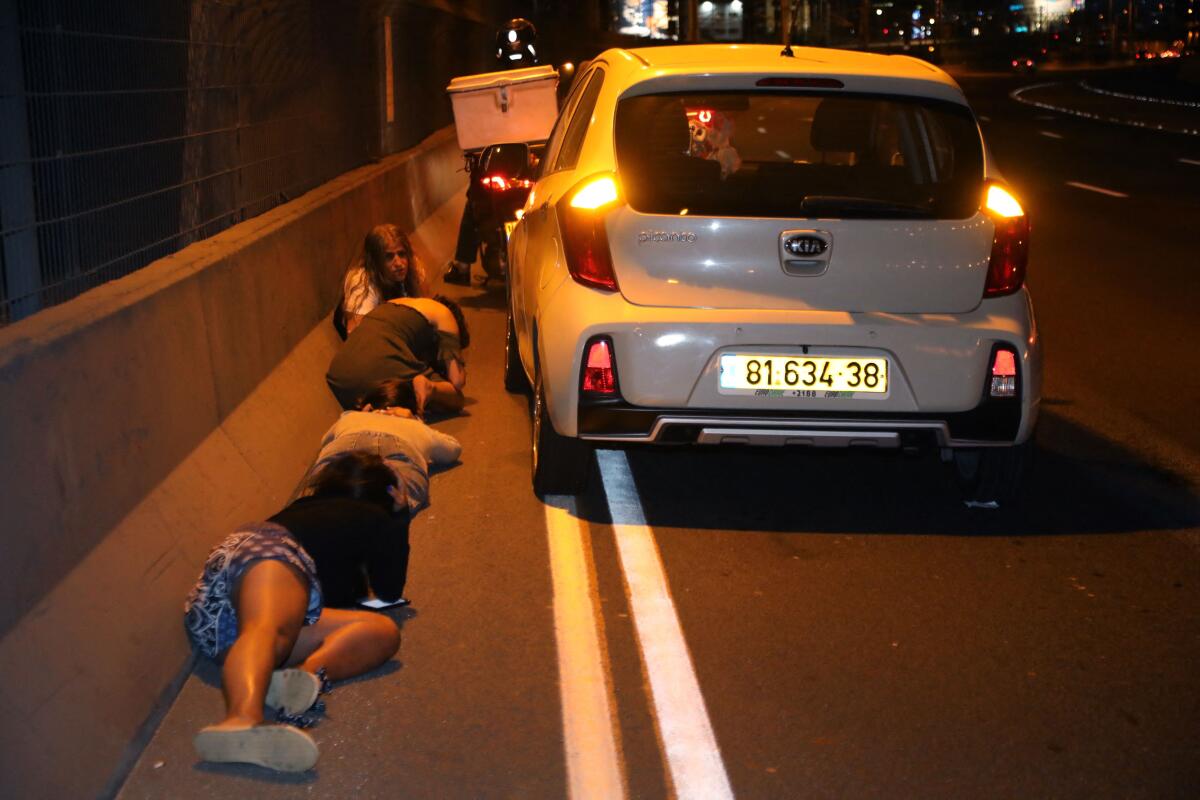 Four people stay low to the ground next to a car on the side of a road