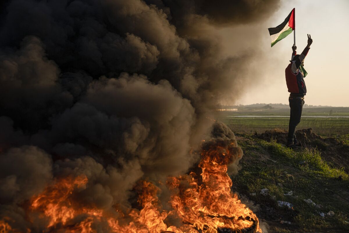 A person holds up a flag next to smoke coming off a pile of burning tires