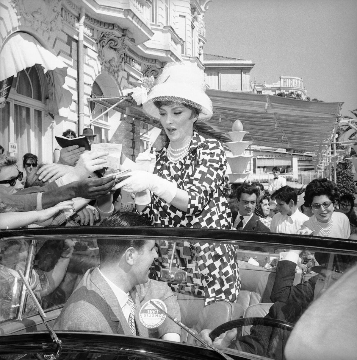 Gina Lollobrigida stands in the back of a car, signing autographs for a smiling crowd.