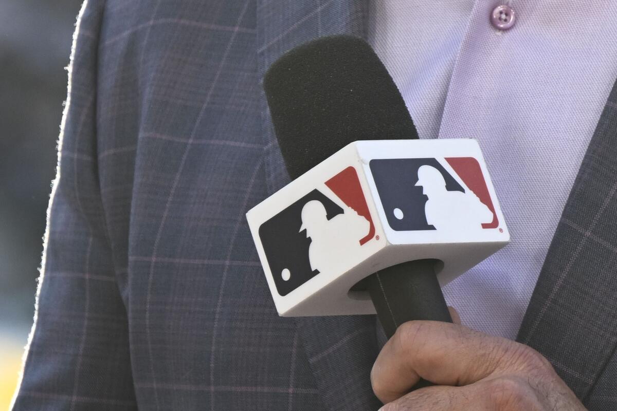 A close-up of an announcer wearing a plaid blazer and lavender shirt while holding a microphone with MLB's silhouette logo