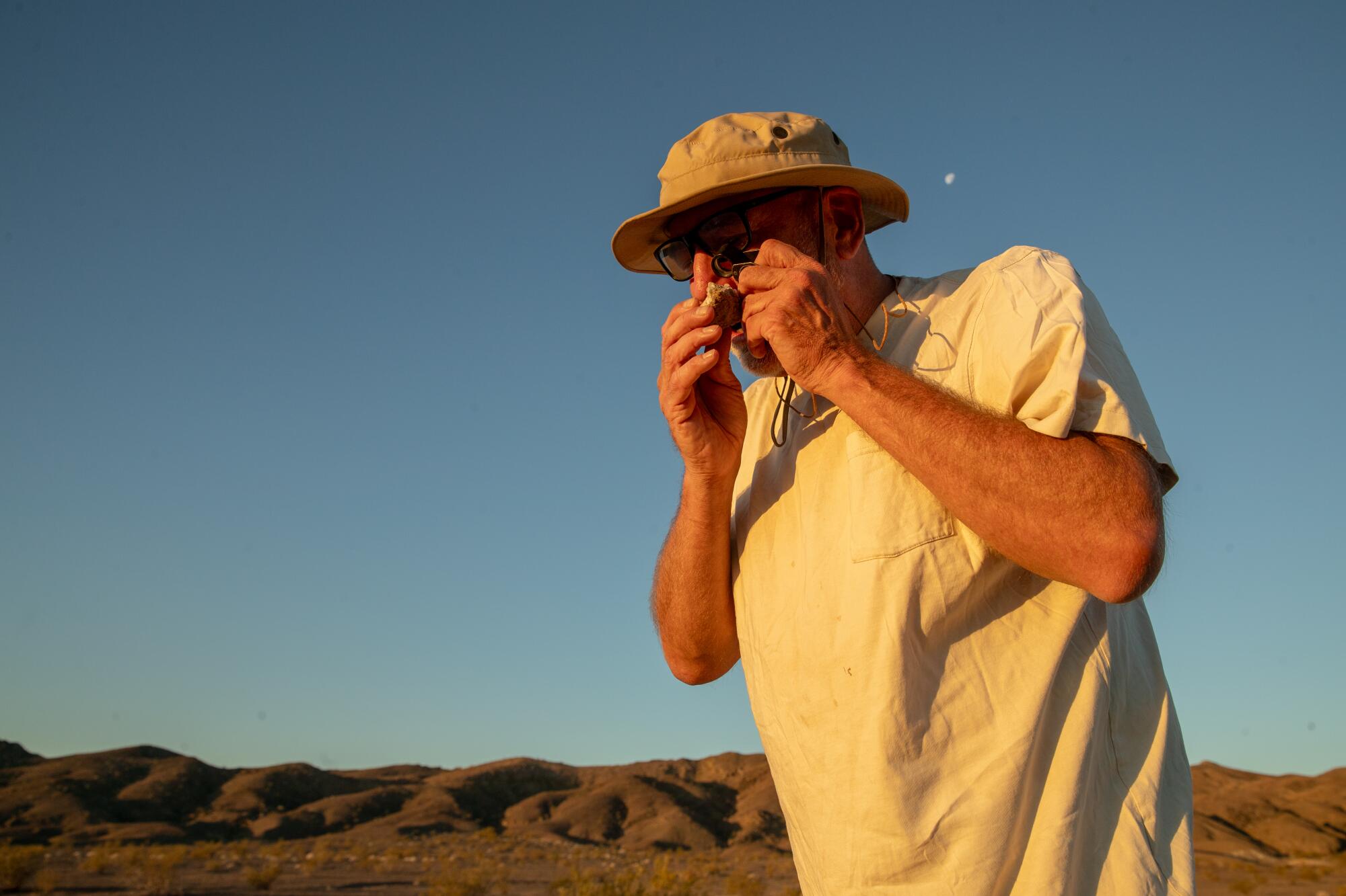 A man uses a magnifying glass to examine a rock in the desert.
