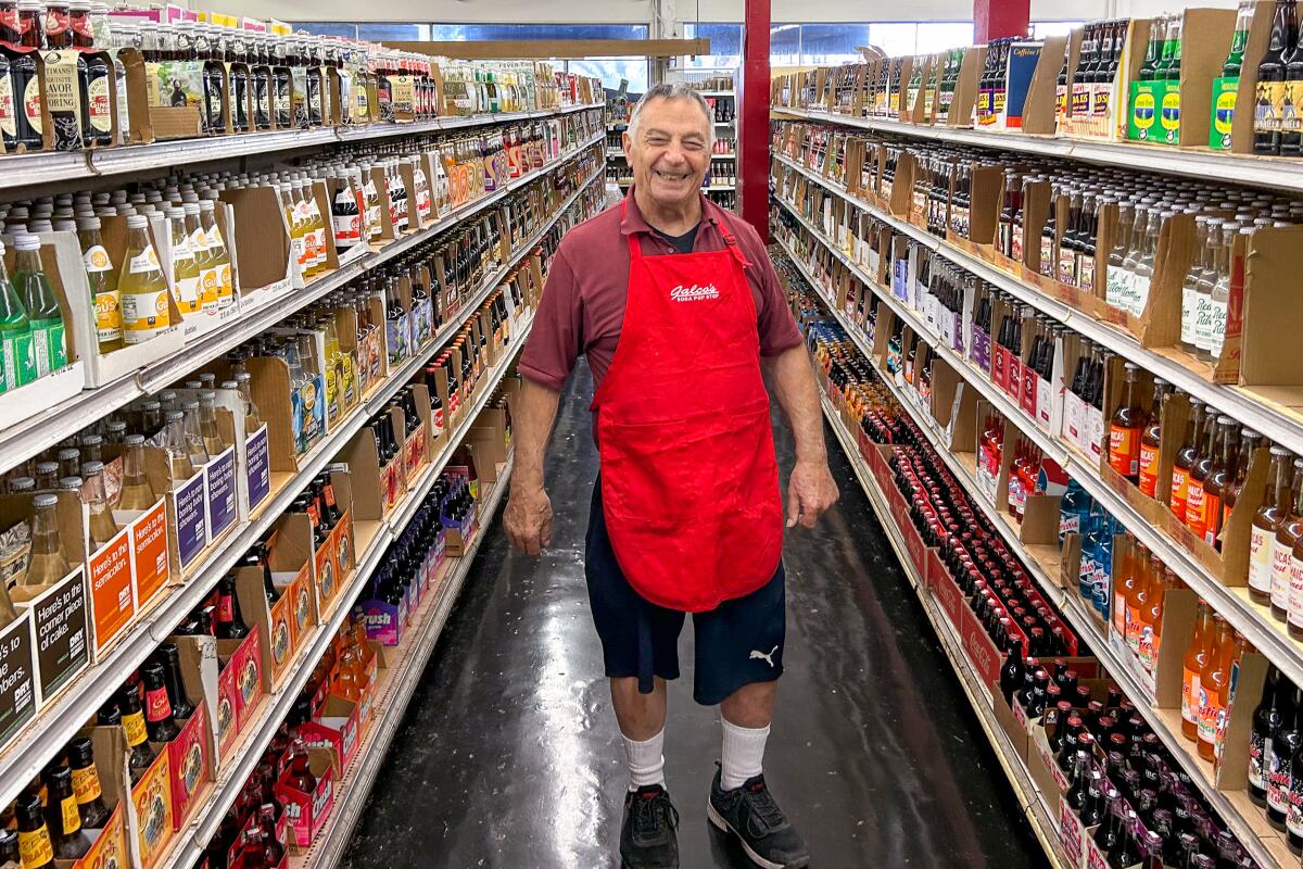 A man wearing a red apron stands in a grocery store aisle between shelves full of bottled drinks.